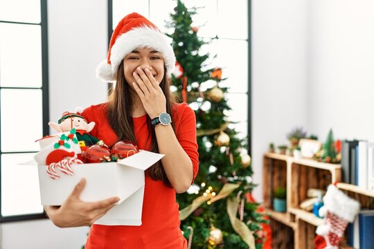 Young brunette woman standing by christmas tree holding decoration laughing and embarrassed giggle covering mouth with hands, gossip and scandal concept