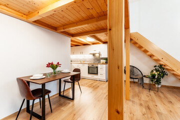 Loft apartment with kitchen, which is under wooden construction forming second floor with staircase. Room features decorative plants, set dining table and regular kitchen appliances.
