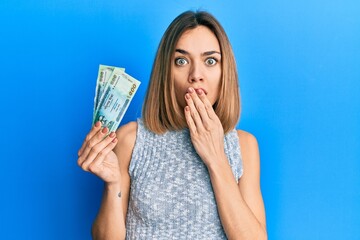 Young caucasian blonde woman holding bangladesh taka banknotes covering mouth with hand, shocked...