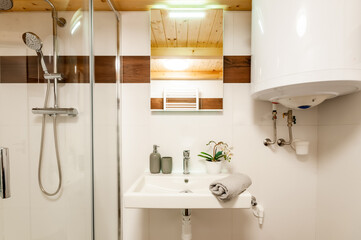 Small modern bathroom with wooden ceiling. In bathroom can be found washbasin, mirror with light and shower in corner. There are also other accessories as soap dispenser, boiler and decorative plant.
