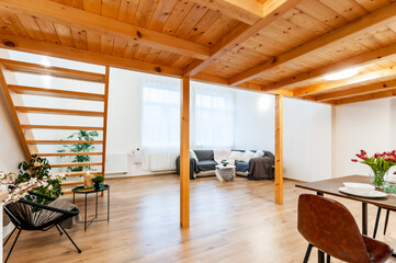 Bright loft apartment with large window, wooden floor and built-in staircase. Room has decorative plants, big corner sofa with few pillows and nice modern chair with small coffee table and small lamp.