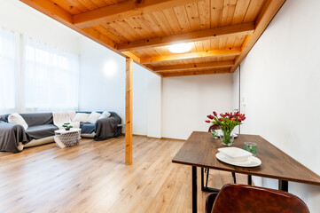 Bright loft apartment with large window, wooden floor and built-in wooden second floor above. Room has decorative coffee table, big corner sofa with pillows, wall mounted light and set dining table.