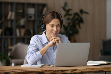 Smiling businesswoman in headphones talking, looking at laptop screen, sitting at work desk,...