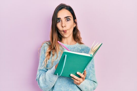 Young hispanic girl reading and writing book making fish face with mouth and squinting eyes, crazy and comical.