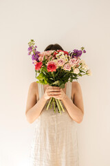 Young beautiful woman in beige washed linen sundress hold bouquet of colourful roses flowers against white wall. Beautiful holiday celebration floral composition
