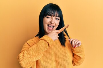 Young hispanic woman styling hair using comb winking looking at the camera with sexy expression,...