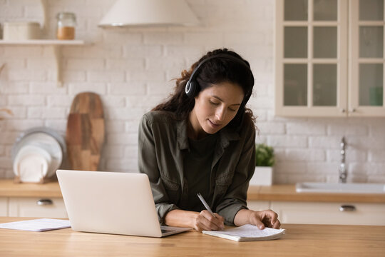 Interested happy beautiful young woman in wireless headphones listening educational online lecture on computer, writing notes in copybook, self educating or improving professional knowledge remotely.