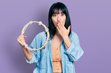 Young hispanic plus size woman playing tambourine covering mouth with hand, shocked and afraid for...