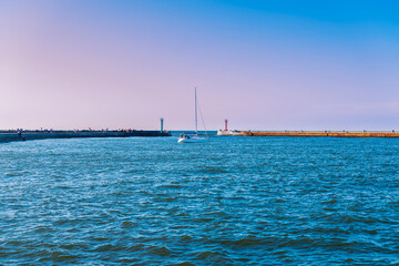 Obraz premium view of the bay in the port, a sailboat entering the port on a sunny day, concrete breakwaters in the background