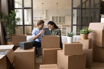 Happy young family couple homeowners unpacking belongings from huge carton boxes, enjoying moving day in modern renovated apartment, enjoying decorating living room together, relocation concept.