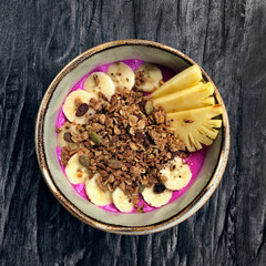 Pitaya breakfast bowl. White rustic black wooden background. Acai superfood smoothie bowl with pineapple slices, banana, dragon fruit smoothie, granola. Overhead, top view, flat lay, copy space.