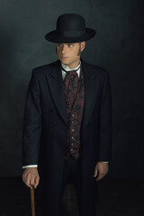 Young man in stylish vintage victorian attire. Standing with a cane in front of a dark gray wall.