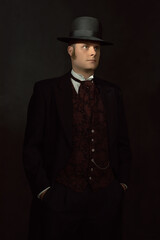 Shadowy portrait of a young man in stylish vintage Victorian attire in front of a dark gray wall.