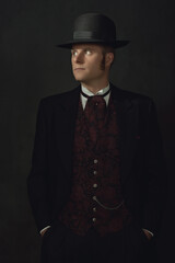 Shadowy portrait of a young man in stylish vintage Victorian attire in front of a dark gray wall.