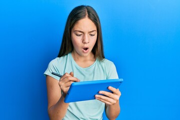 Beautiful brunette little girl using touchpad drawing on screen in shock face, looking skeptical and sarcastic, surprised with open mouth