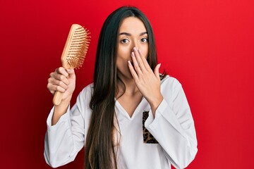 Young hispanic girl holding hairbrush covering mouth with hand, shocked and afraid for mistake....