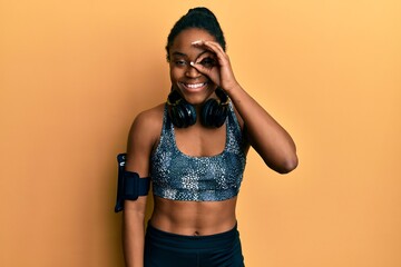 African american woman with braided hair wearing sportswear and arm band doing ok gesture with hand smiling, eye looking through fingers with happy face.