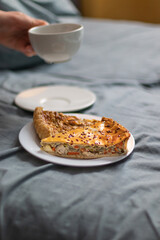 Two slices of homemade quiche pie on a white plate and a cup of aromatic chamericano coffee on the bed at home