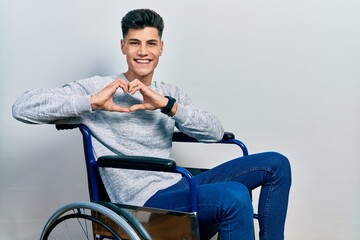 Young hispanic man sitting on wheelchair smiling in love doing heart symbol shape with hands. romantic concept.