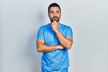 Handsome hispanic man with beard wearing blue male nurse uniform with hand on chin thinking about...