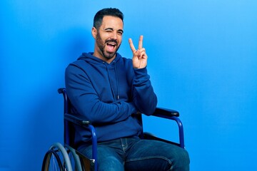 Handsome hispanic man with beard sitting on wheelchair smiling with happy face winking at the camera doing victory sign. number two.