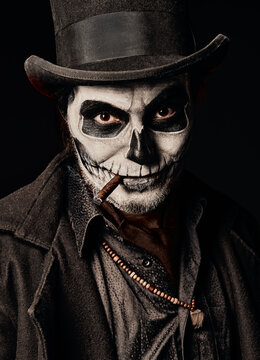 Young man in image of Baron Samedi, the Voodoo deity. Baron Saturday dressed in black coat and top hat smoking cigar, close-up portrait. Day of the Dead (and Halloween) theme