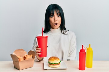 Young brunette woman with bangs eating a tasty classic burger with ketchup and mustard scared and amazed with open mouth for surprise, disbelief face