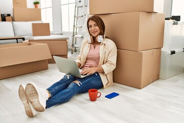 Middle age caucasian woman using laptop and headphones sitting on the floor at new home.