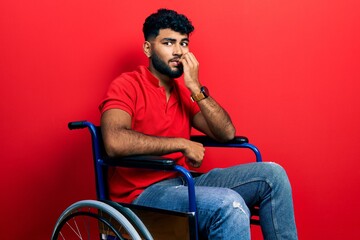 Arab man with beard sitting on wheelchair looking stressed and nervous with hands on mouth biting...