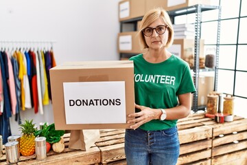 Middle age blonde woman wearing volunteer t shirt holding donations box thinking attitude and sober...