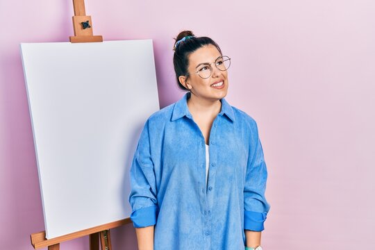 Young hispanic woman standing by painter easel stand looking away to side with smile on face, natural expression. laughing confident.