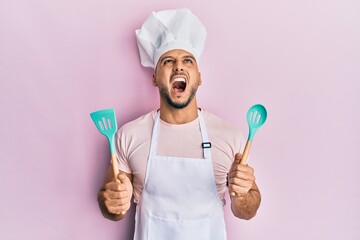 Young arab man wearing professional cook apron and hat holding spoon angry and mad screaming...