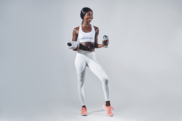 Happy young African woman in sports clothing holding bottle with water and exercise mat
