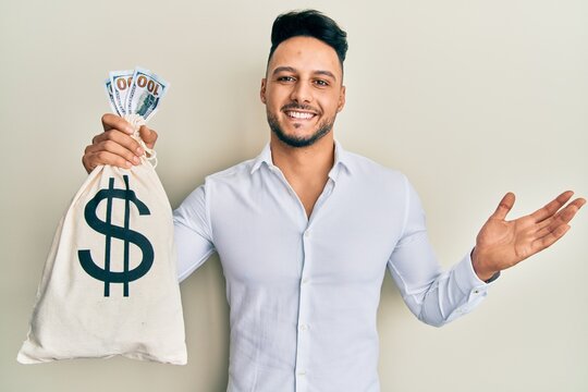 Young arab man holding dollars bag celebrating achievement with happy smile and winner expression with raised hand