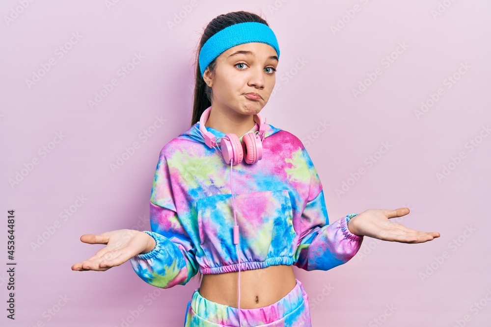 Sticker young brunette girl wearing gym clothes and headphones clueless and confused expression with arms an