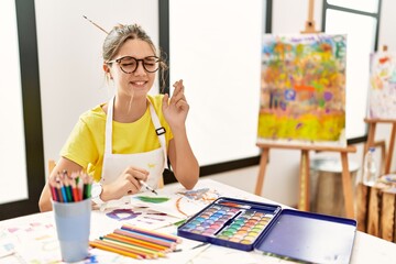 Young brunette teenager at art studio gesturing finger crossed smiling with hope and eyes closed....