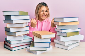 Young caucasian woman sitting on the table with books very happy and excited doing winner gesture with arms raised, smiling and screaming for success. celebration concept.