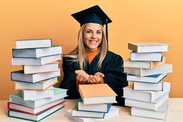 Young caucasian woman wearing graduation ceremony robe sitting on the table smiling with hands palms together receiving or giving gesture. hold and protection