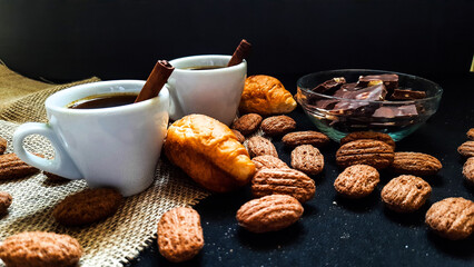 Two small white coffee mugs with cinnamon, croissants and chocolate on a dark background