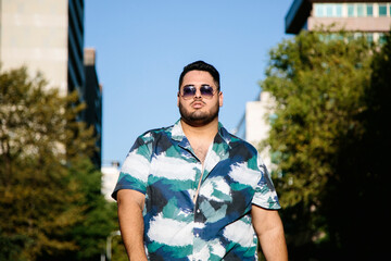 Fat latin man with sunglasses, walking, with a serious expression. With a street in the background....