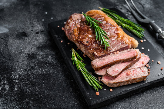Cut roasted new york strip beef meat steak or striploin on a marble board. Black background. Top view. Copy space
