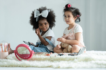 Two little girls friends, African and Caucasian, playing cosmetics together and use pink makeup...