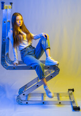 girl in full growth. A model in white sneakers, a T-shirt and blue jeans is sitting on the transforming stairs. multi-colored light on the background and on the face. blue and yellow background