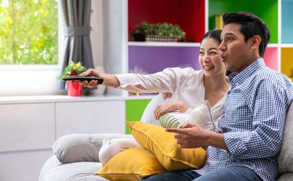 Happy asian parents sit on comfortable couch in living room watch TV enjoy home weekend enjoying movie