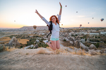 Happy girl jump with a backpack. Posing against the backdrop of a valley with mountains and balloons at sunrise. Entertainment, tourism an vacation. Travel tour. Goreme, Cappadocia, Turkey.