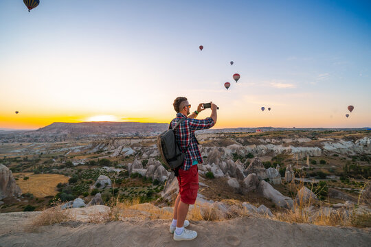 Man with backpack takes a picture, photo on smartphone. Tourist attraction balloon flight on background. Entertainment, tourism an vacation. Travel tour. Goreme, Cappadocia, Turkey.