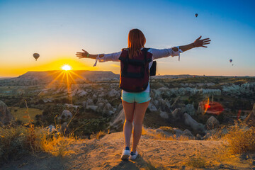 Girl with arms spread apart posing. Valley with mountains and balloons at sunrise. Back view. Entertainment, tourism an vacation. Travel tour. Goreme, Cappadocia, Turkey.