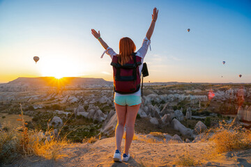 Girl with arms spread apart posing against the backgrond of a valley with mountains and balloons at sunrise. Back view. Entertainment, tourism an vacation. Travel tour. Goreme, Cappadocia, Turkey.
