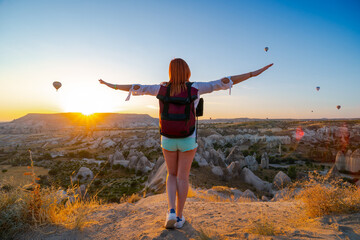 Girl with arms spread apart posing against the backgrond of a valley with mountains and balloons at sunrise. Back view. Entertainment, tourism an vacation. Travel tour. Goreme, Cappadocia, Turkey.