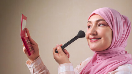 Woman in hijab applying makeup with a brush while looking in the mirror.